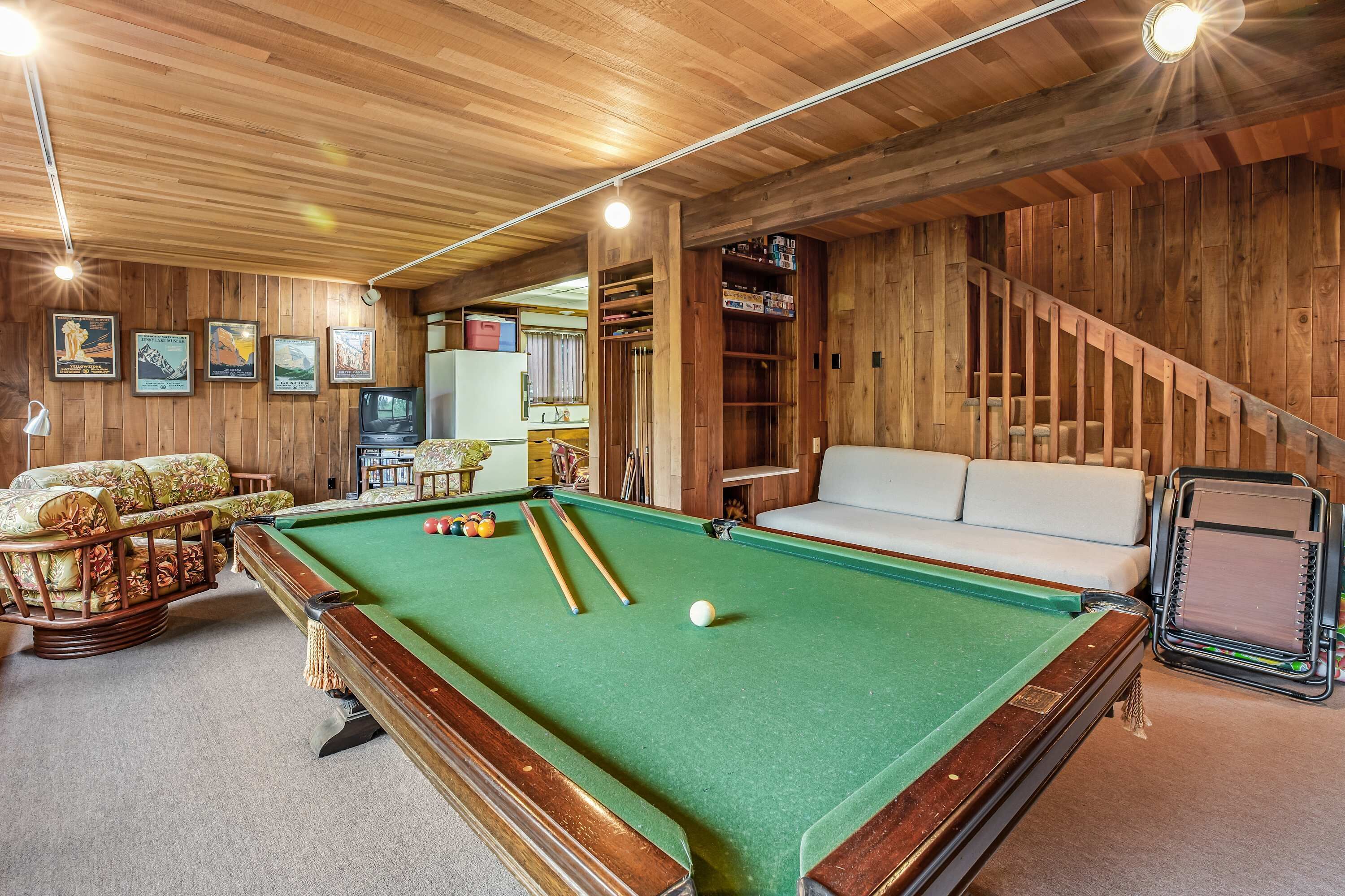 A pool table with cues and balls, a sofa, and a staircase in a wooden room.