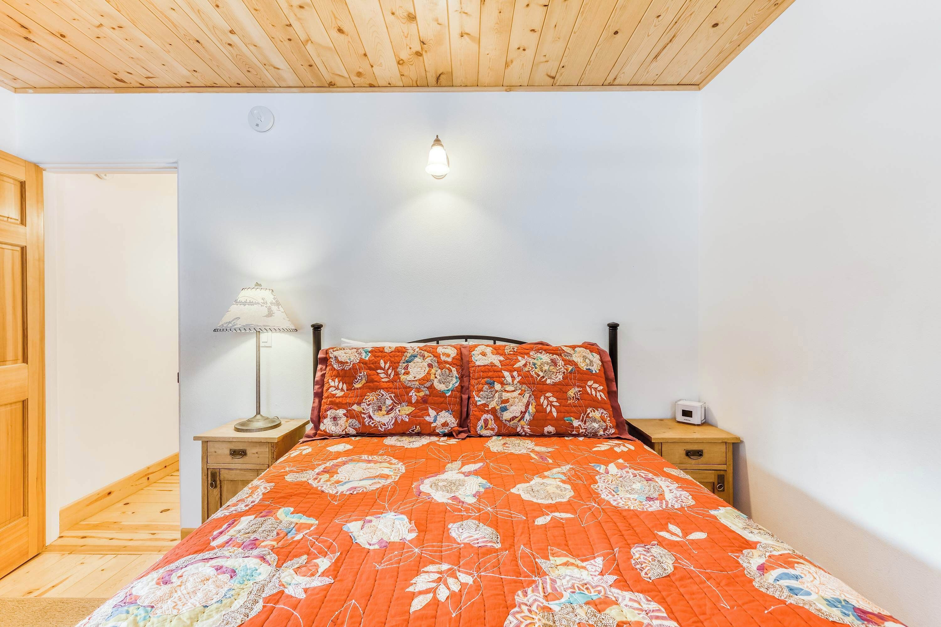 Une chambre avec un plafond en bois, un lit recouvert d’une literie à motifs floraux, une lampe de chevet et une table de chevet avec une radio.