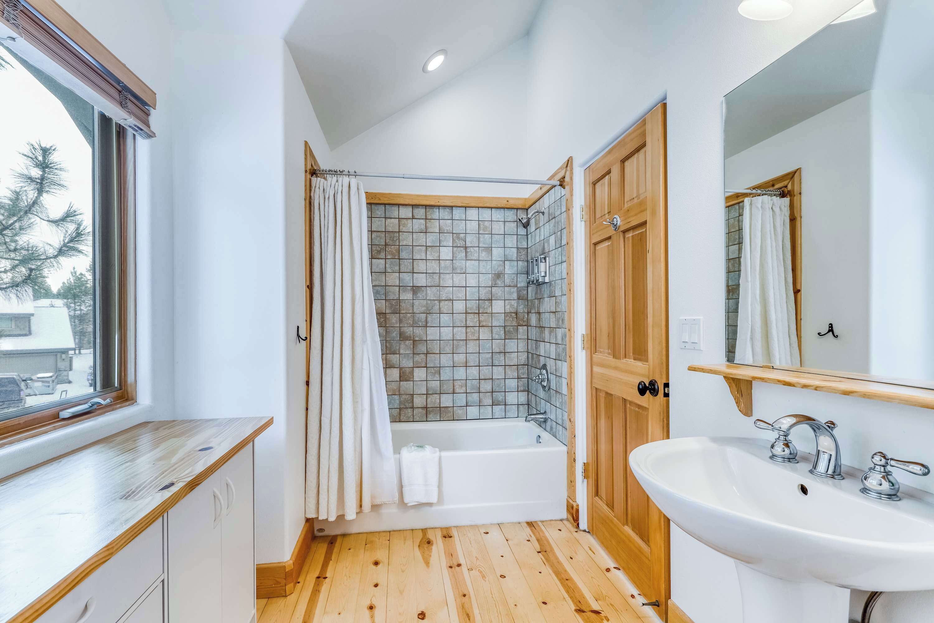 A bathroom with a wooden floor, a white bathtub, a white sink, a wooden door, and a window with a view of trees.