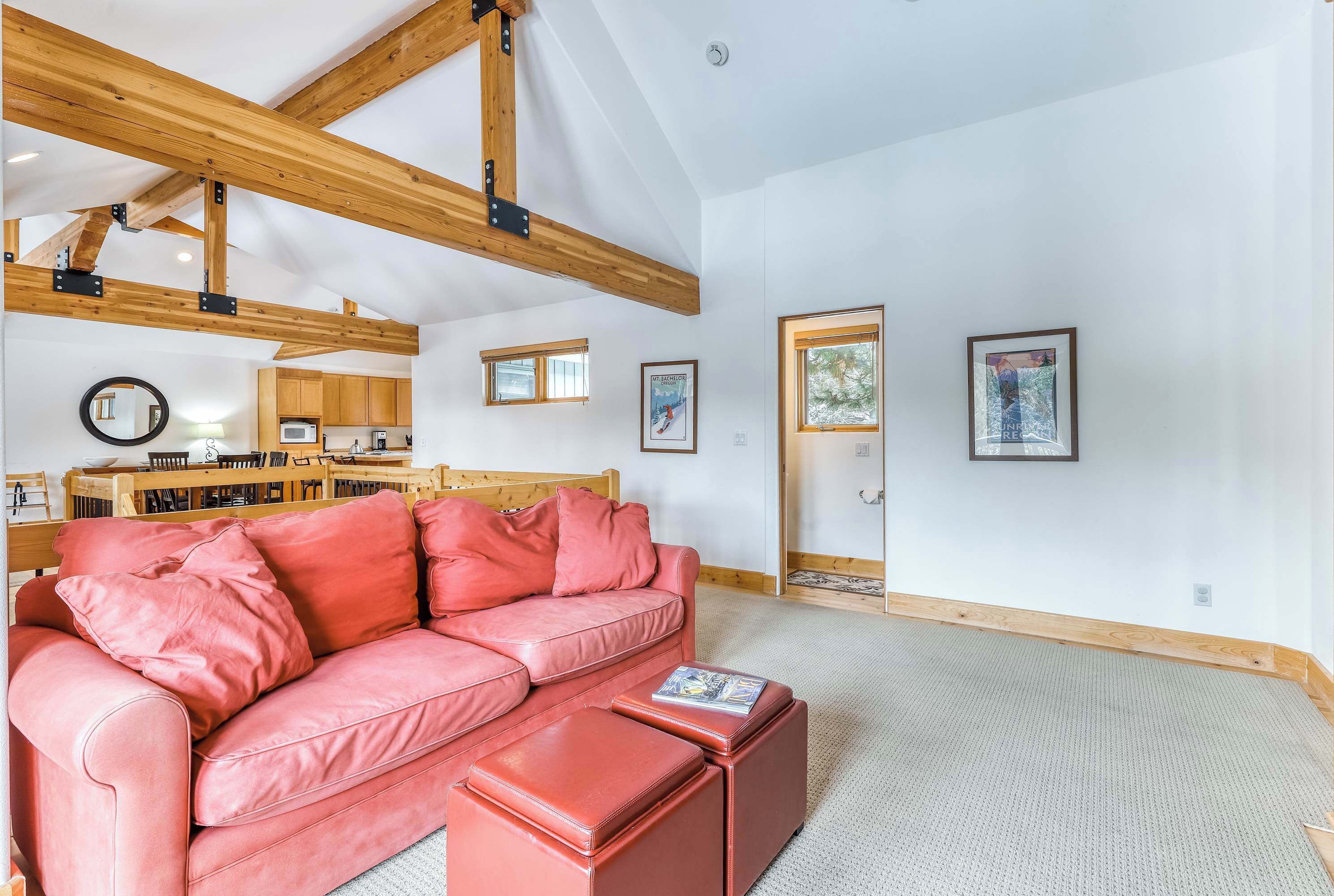 A spacious living room with a red leather sofa, wooden beams, a kitchen in the background, and framed pictures on the wall.