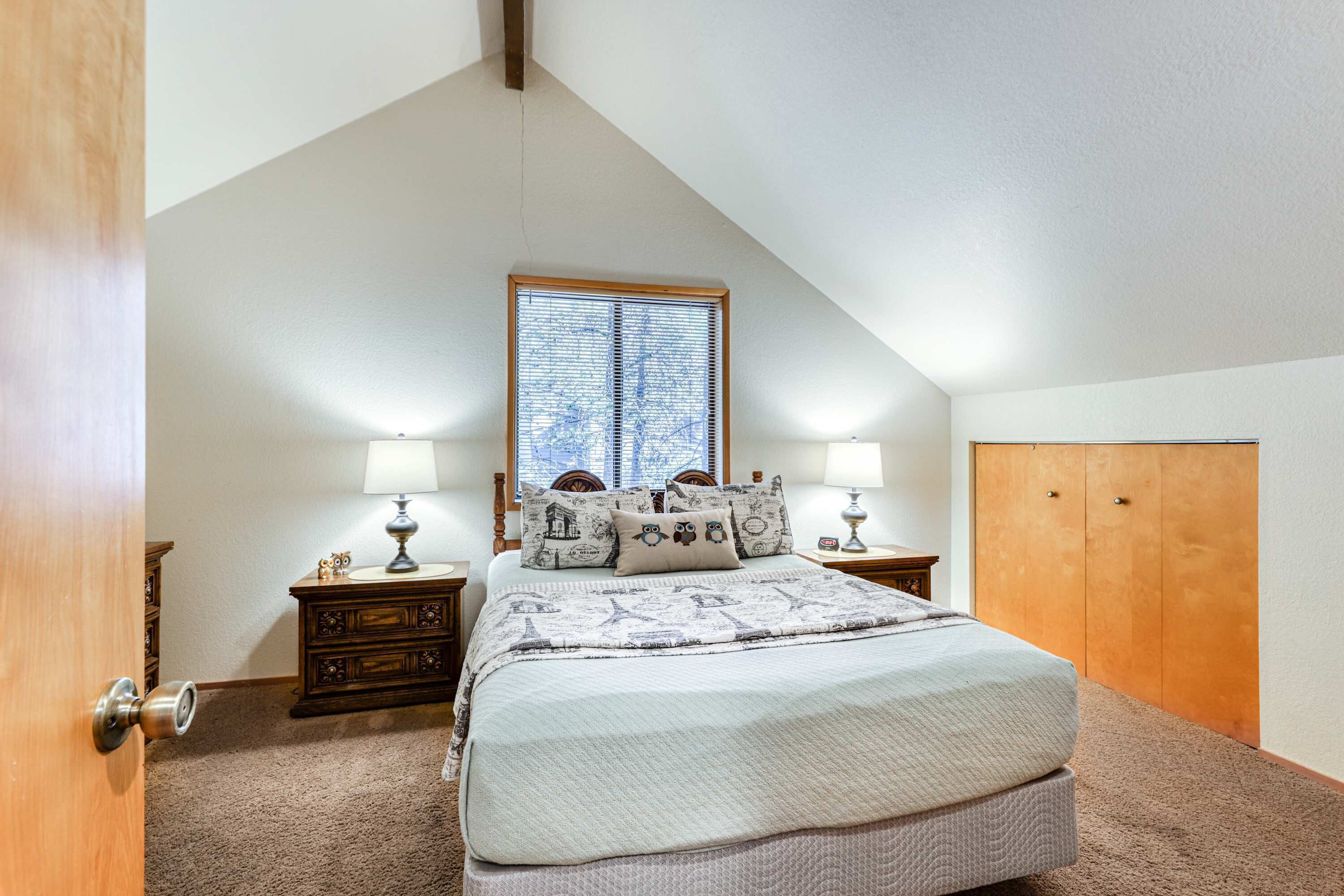 A bedroom with a sloped ceiling, a large window with blinds, a bed with a quilt, two bedside tables with lamps, and a wooden wardrobe.