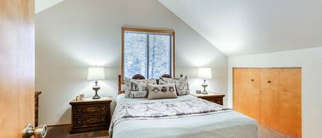 A bedroom with a sloped ceiling, a large window with blinds, a bed with a quilt, two bedside tables with lamps, and a wooden wardrobe.