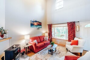 A living room with a red sofa, white armchair, and a coffee table. There is a window with curtains, a fireplace, and a TV.
