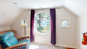 A room with a sloped ceiling, a window with purple curtains, a small table, and a blue chair. - Bay & Harbor View Home with High-Speed WiFi, Private Washer/Dryer, & Board Games (Newport)