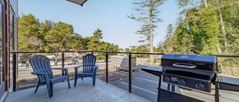 A balcony with a grill, two Adirondack chairs, and a view of trees and a parking lot.
