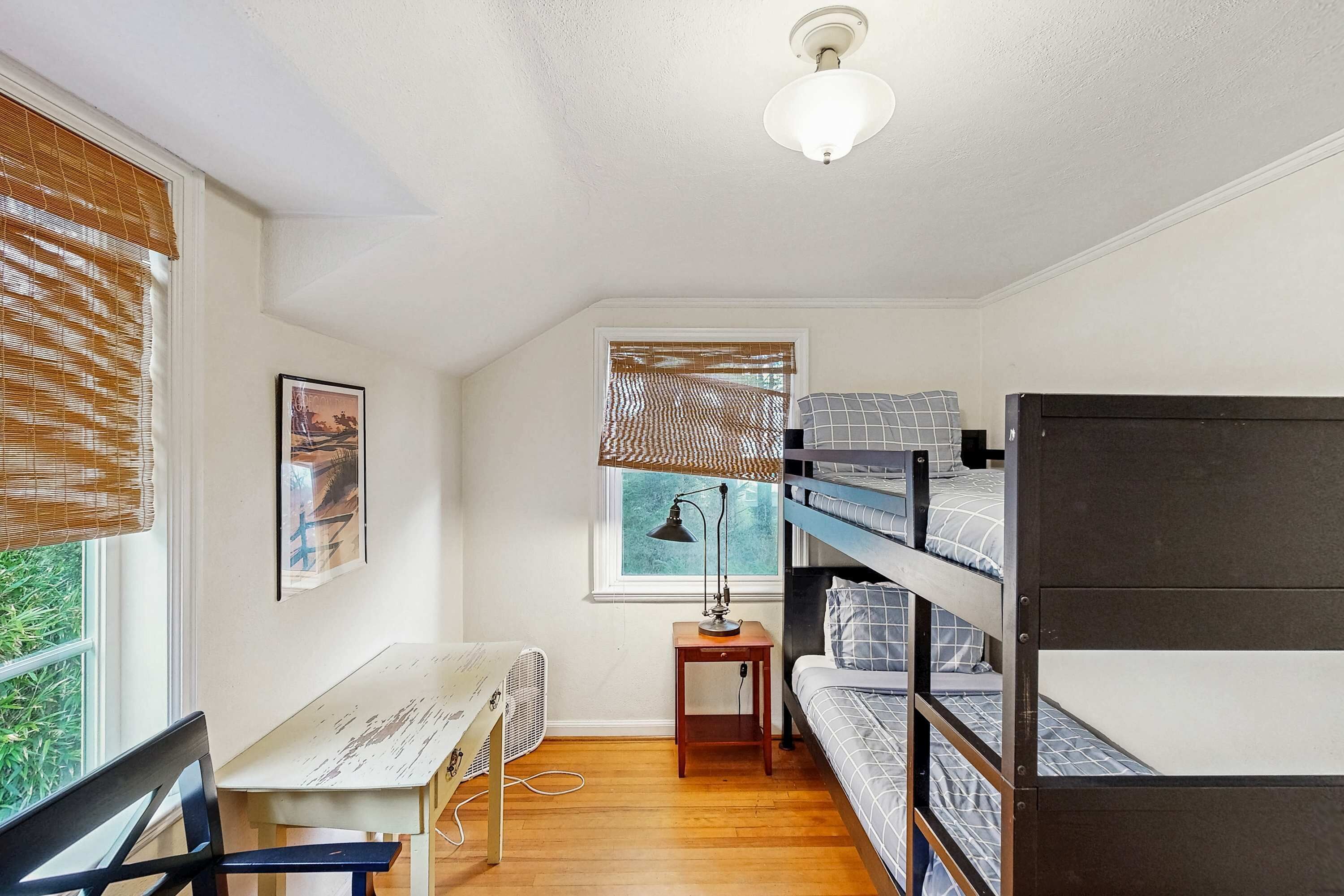 A compact room with bunk beds, a desk, a chair, a standing fan, and a window with blinds.