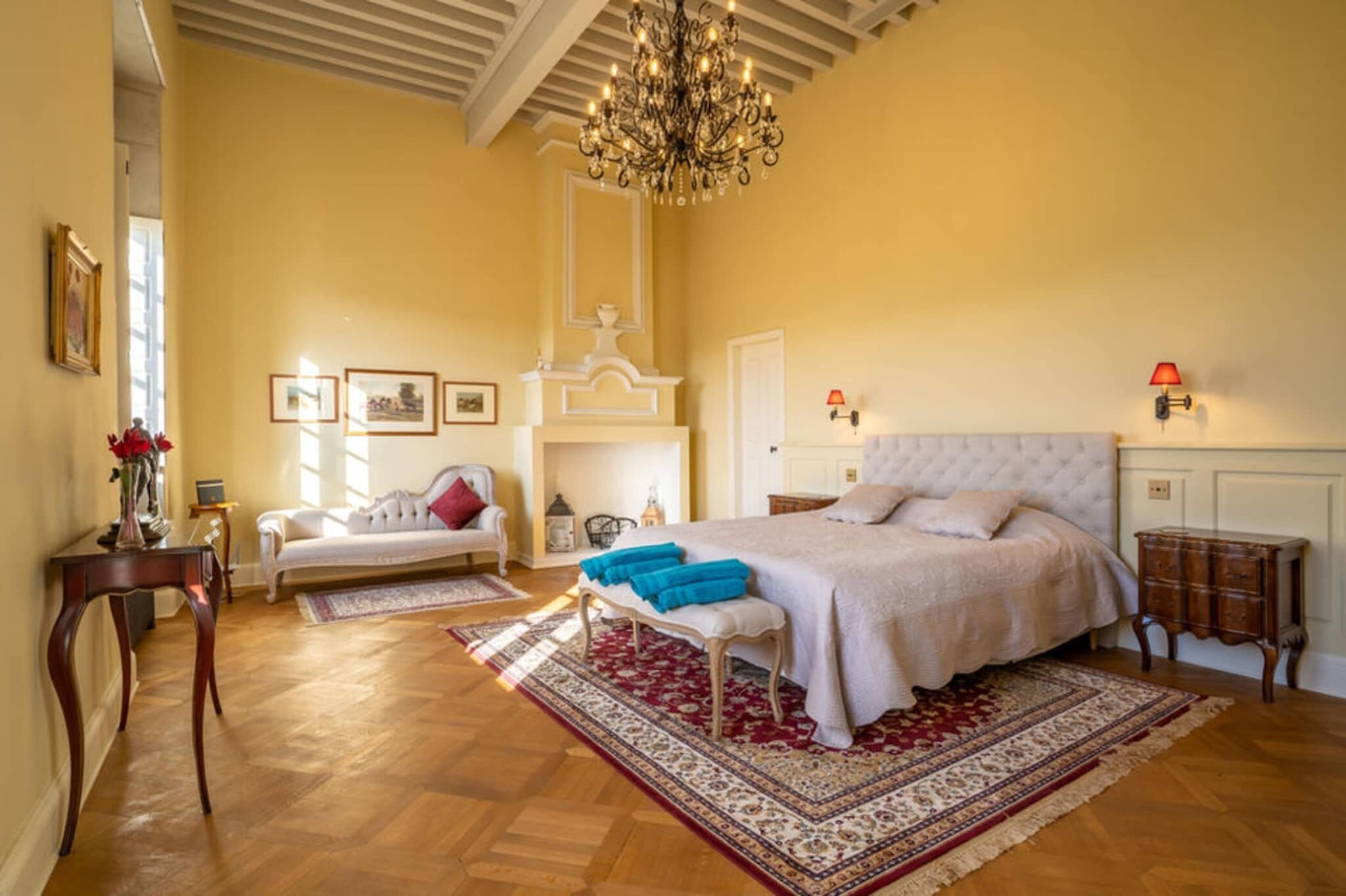 A bedroom with a large bed, a chandelier, a white sofa, a small table, and a red rug.