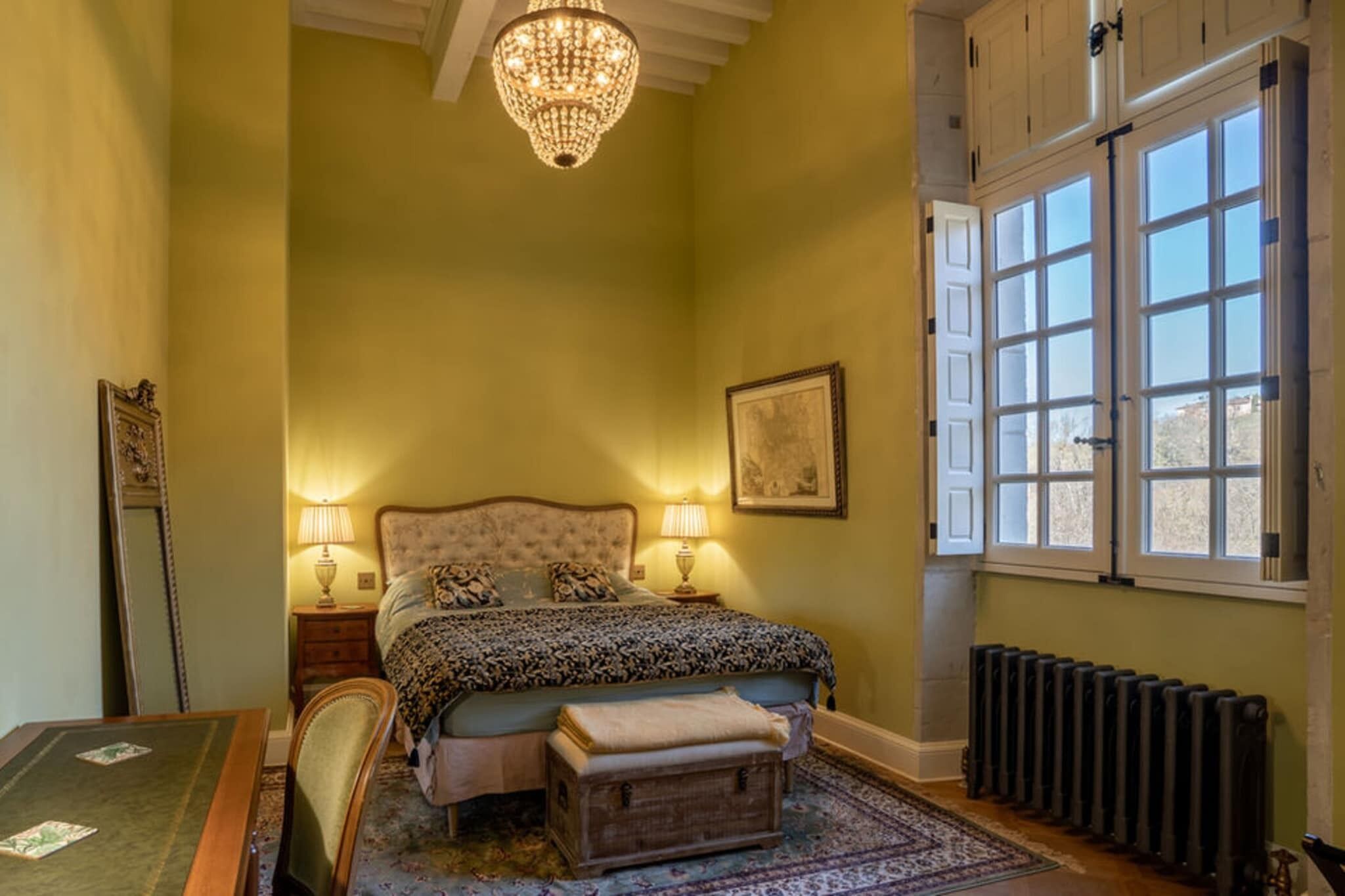 Une chambre à coucher avec un grand lit, deux lampes de chevet, un lustre, un radiateur et une fenêtre donnant sur un beau paysage.