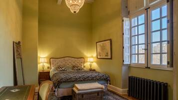 A bedroom with a large bed, two bedside lamps, a chandelier, a radiator, and a window with a view.