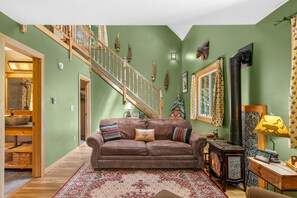 A living room with a wooden staircase, a brown sofa, a patterned rug, and a wood-burning stove.
