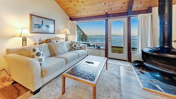 A cozy living room with a sofa, a coffee table, a fireplace, and a large window overlooking a scenic view. - Waterfront Condo with Private Deck, Bay Views, Pool, Hot Tub, & Sauna (Newport)