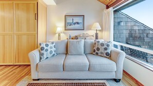 A room with a wooden ceiling, a grey sofa with patterned cushions, a framed picture on the wall, and a window with a view of a building. - Waterfront Condo with Private Deck, Bay Views, Pool, Hot Tub, & Sauna (Newport)