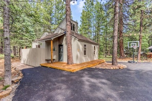 A cabin with a wooden deck, a basketball hoop, and a paved driveway surrounded by trees. - Cozy home with shared pool, sauna, golf, private hot tub & deck - dog-friendly (Black Butte Ranch)