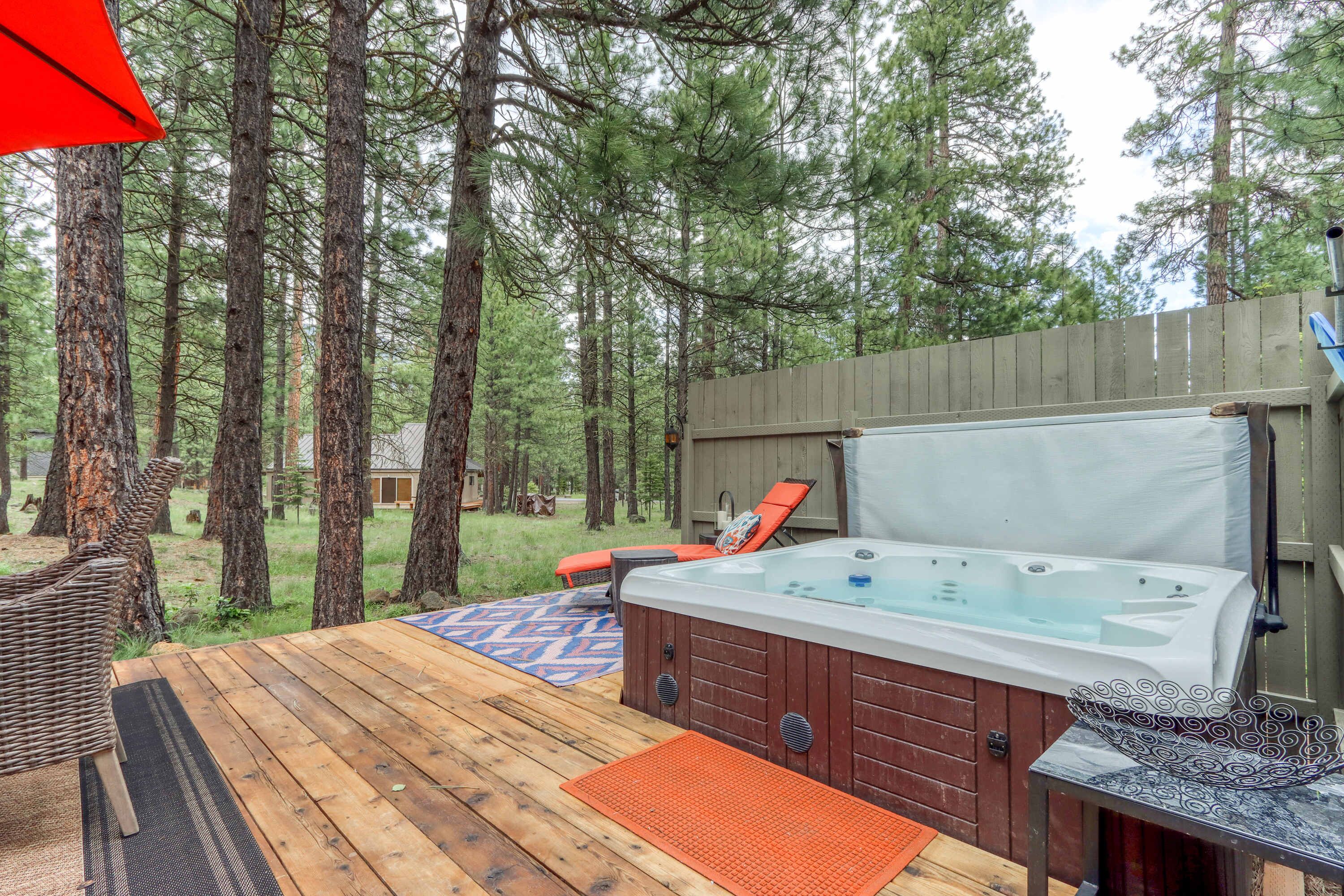 A hot tub on a wooden deck surrounded by trees, with a wooden fence and a small building in the background.
