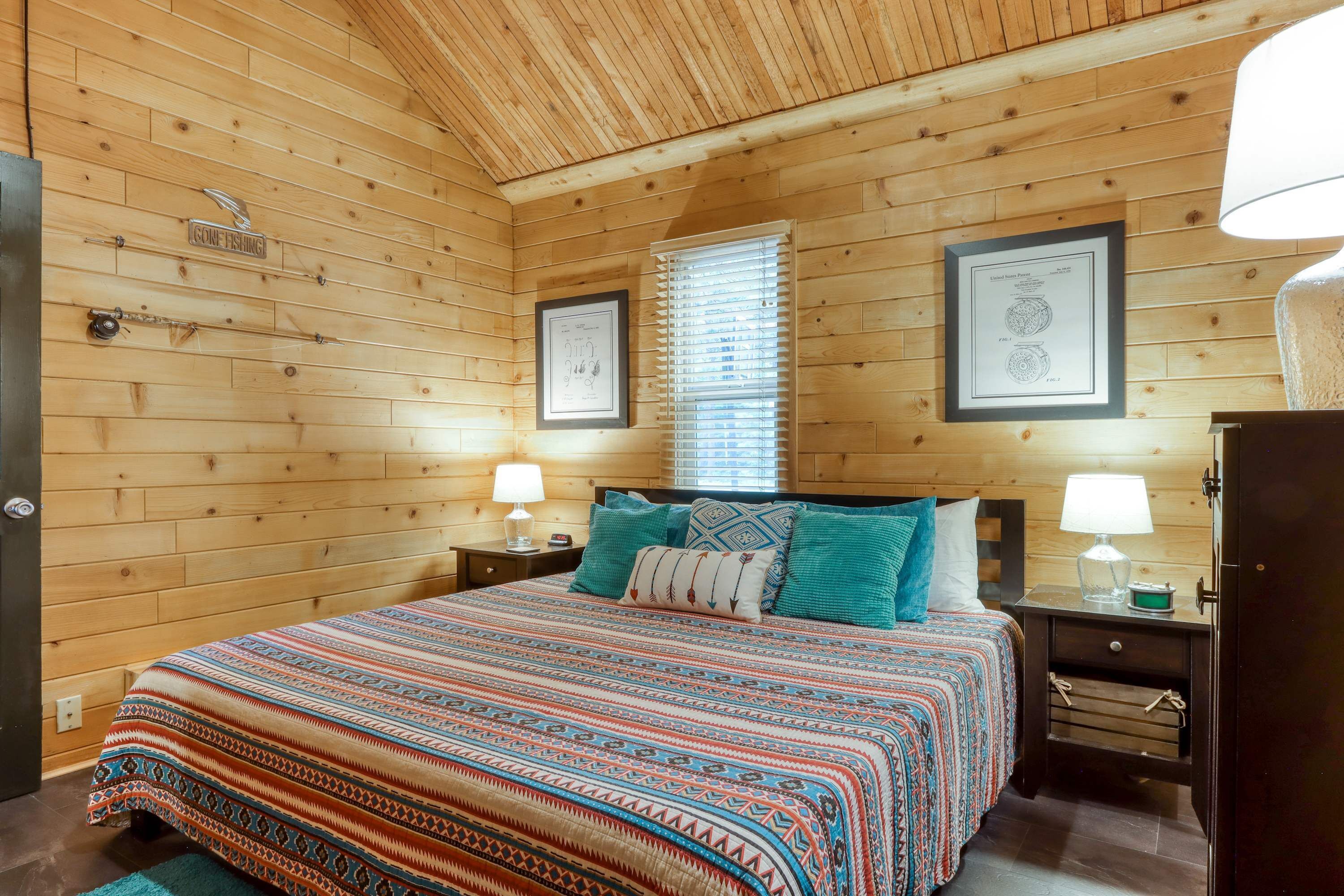 A wooden cabin bedroom with a bed, two nightstands, a window with blinds, and framed certificates on the wall.