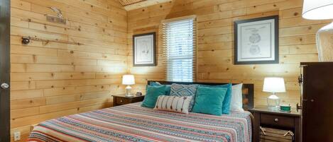 A wooden cabin bedroom with a bed, two nightstands, a window with blinds, and framed certificates on the wall.