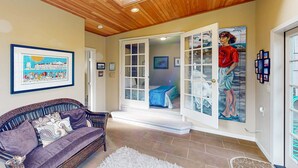 Una habitación con un sofá de mimbre, un cuadro enmarcado y una puerta que da a otra habitación con una cama y un cuadro.