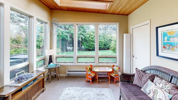 Una habitación luminosa con ventanas amplias, techo de madera, sofá, mesa con sillas y un radiador.
