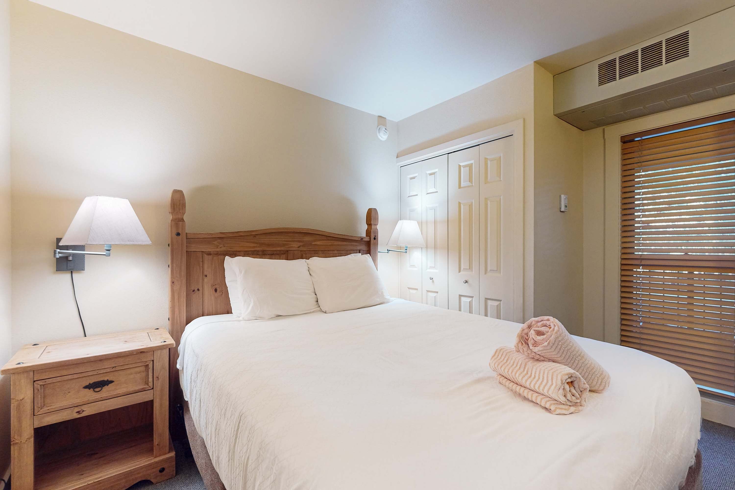 Une chambre à coucher avec une tête de lit en bois, un lit avec une literie blanche, deux lampes de chevet, une table de chevet, une armoire et une fenêtre avec des stores.