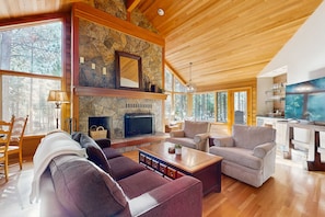 A spacious living room with a stone fireplace, wooden ceiling, and large windows. - Tree-lined home with private hot tub, 7 pools, 14 tennis courts, golf, gym, AC (Black Butte Ranch)