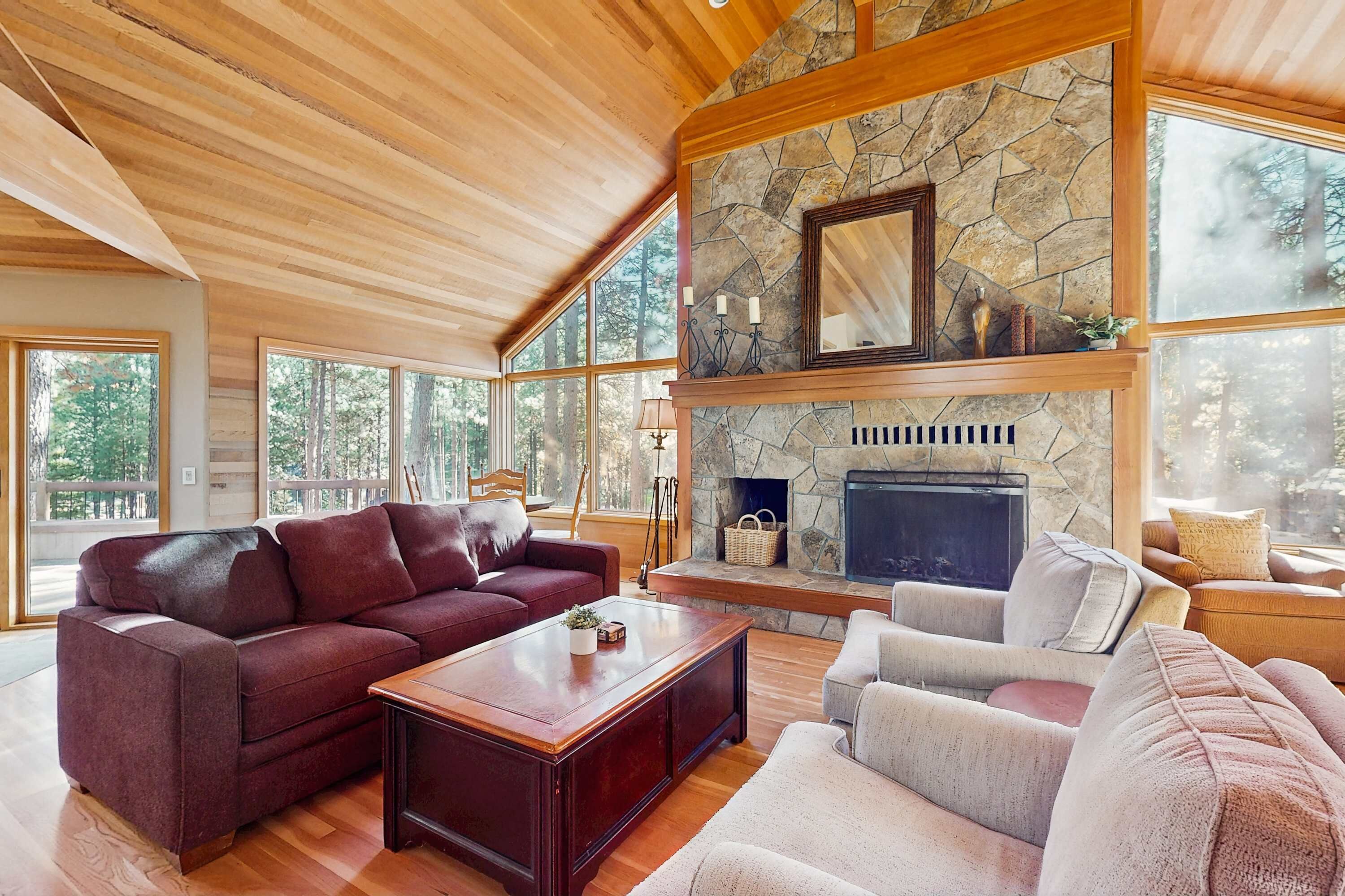 A spacious living room with a stone fireplace, wooden ceiling, and large windows.