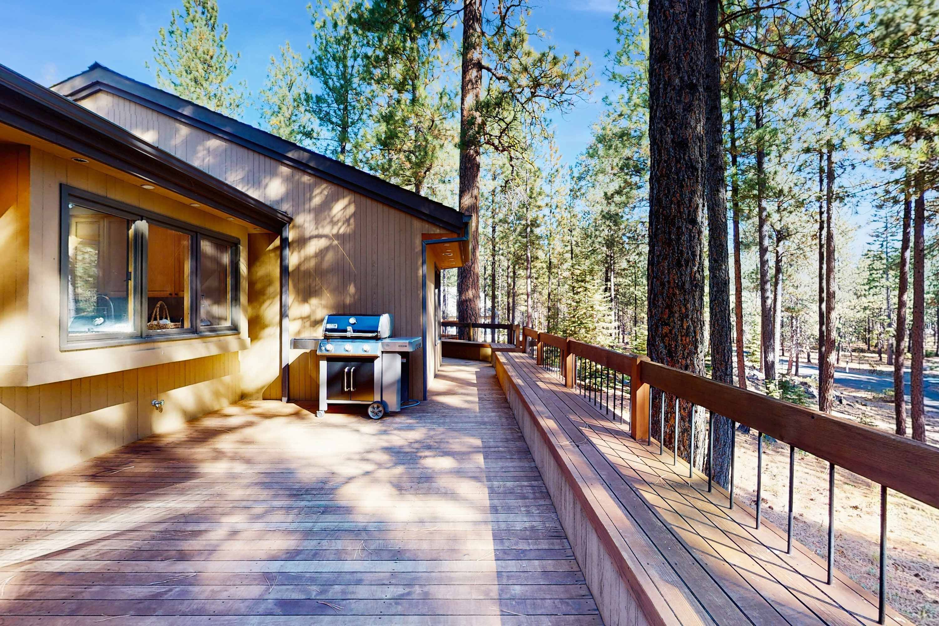 A wooden deck with a grill, a wooden building with large windows, and a forested backdrop.