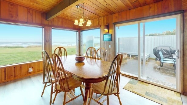 A dining area with a wooden table and chairs, a television, and large windows with a view of the outdoors. - Oceanfront, Dog-Friendly Home with Fireplace, Ocean View, Beach Access & WiFi (Yachats)