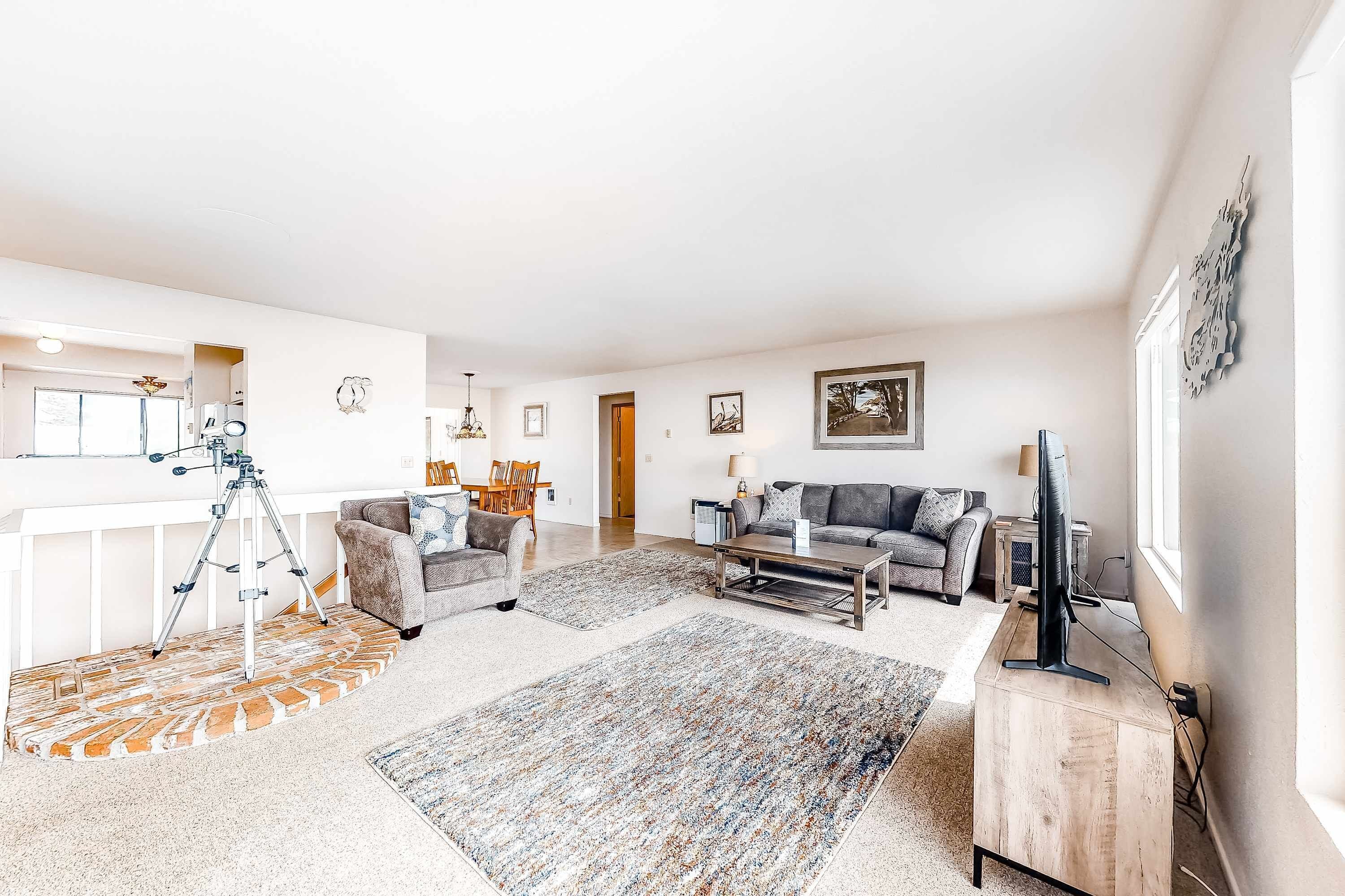 A spacious living room with a sofa, coffee table, and a television. There is a dining area with chairs and a table, and a kitchen area with a fridge and a microwave.
