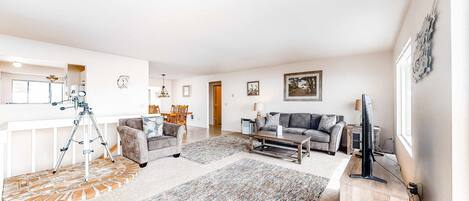 A spacious living room with a sofa, coffee table, and a television. There is a dining area with chairs and a table, and a kitchen area with a fridge and a microwave.