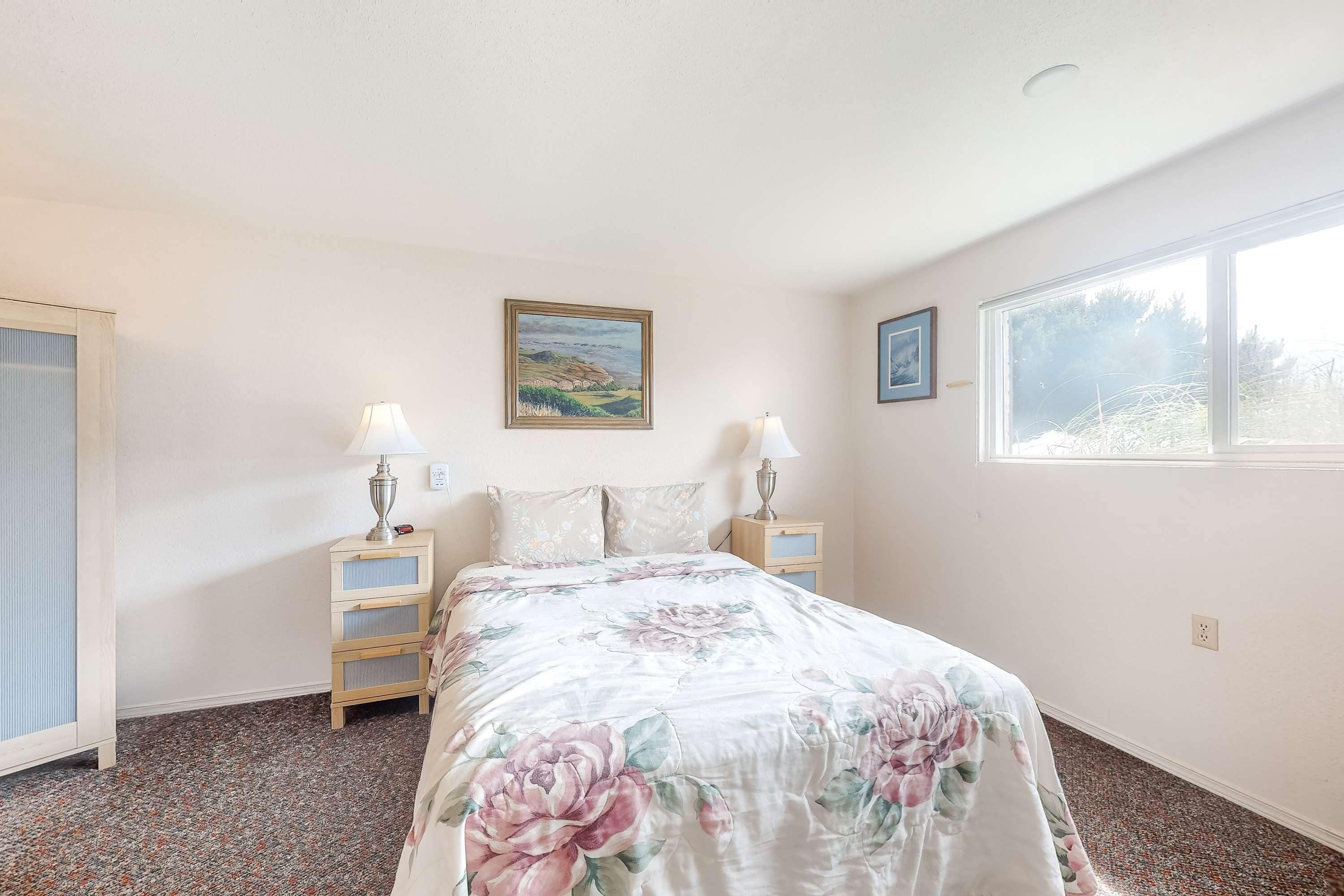 A bedroom with a floral bedspread, two bedside lamps, a nightstand, a framed picture on the wall, and a window with a view.