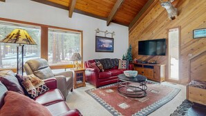 A cozy living room with a leather sofa, a red couch, a round coffee table, a mounted deer head, and a TV. - Rustic-chic home with patio, pools, hot tubs, & fitness center (Black Butte Ranch)