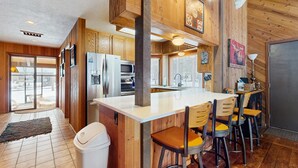 A kitchen with wooden cabinets, a central island with bar stools, a refrigerator, and a microwave. - Rustic-chic home with patio, pools, hot tubs, & fitness center (Black Butte Ranch)