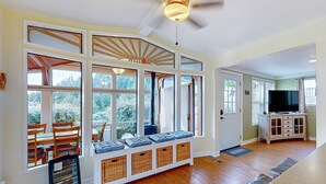 A room with a large bay window, a ceiling fan, a wooden bench with cushions, and a television on a cabinet. - Oceanfront home with private hot tub, walls of glass & spectacular view (Brookings)