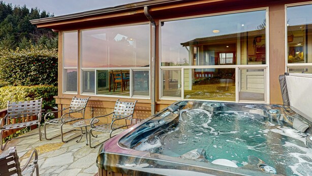 A hot tub with stone tiles, a wooden deck, and a view of a house with large windows. - Oceanfront home with private hot tub, walls of glass & spectacular view (Brookings)