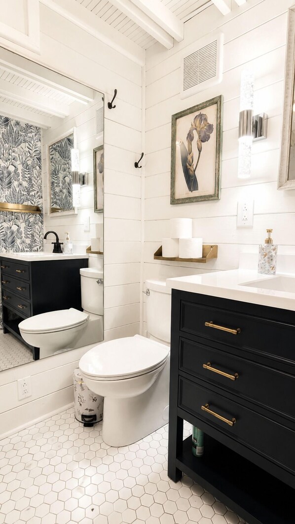 A bathroom with two toilets, a black vanity with gold handles, a white sink, and a mirror.