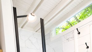 Une douche fermée par des parois de verre, avec des carreaux de marbre blanc, une étagère en bois et une vue sur la verdure à l’extérieur.