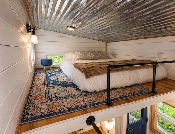 Dormitorio tipo loft con cama, un reposapiés azul, alfombra estampada y vista a zonas verdes.