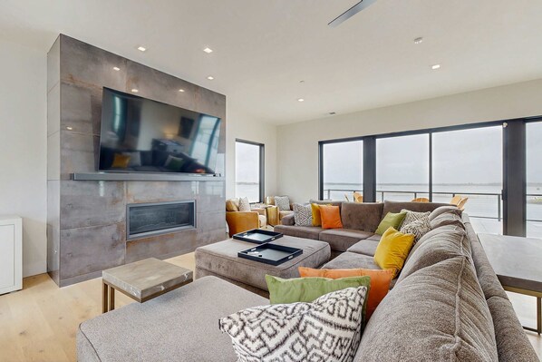 A modern living room with a large flat-screen TV mounted on a stone wall, a sectional sofa with multiple cushions, a coffee table, and large windows offering a view of the outdoors.