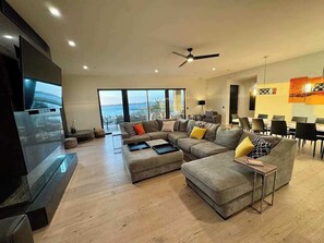 A spacious living room with a large flat-screen TV, a sectional sofa, and a dining area with chairs and tables.