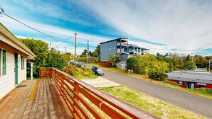 A wooden deck with a red railing, a road with cars, and a modern two-story building with a balcony. - Dog-friendly duplex in the Heart of Nye beach near the beach & downtown (Newport)