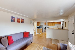 A living room with a sofa and a coffee table, a kitchen with wooden cabinets, and a hallway leading to another room.