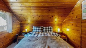 Ein Holzhütten-Zimmer mit einem Bett, zwei Nachttischen, einem fenster mit Vorhängen und einer Wandleuchte.