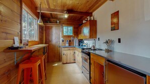 Eine Holzhütte mit einer Küchenzeile, einschließlich Holzausziehtisch, Herd und Spüle. Holzschemeln und ein Fenster mit Blick auf Bäume.