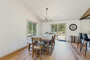 A dining area with a wooden table and chairs, a chandelier, and large windows. - Remodeled Luxury Home with private hot tub, patio-balcony, & SHARC Passes (Sunriver)