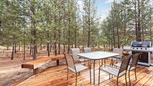 Une terrasse extérieure avec une table, des chaises, un barbecue et un banc, le tout entouré d’arbres.