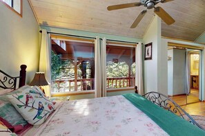 Ein Schlafzimmer mit Bett, Deckenventilator und großen Fenstern mit Blick auf Bäume.
