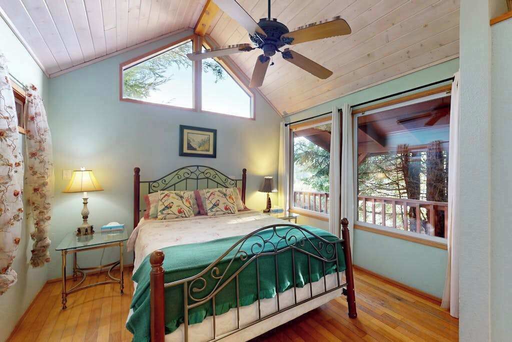 A bedroom with a bed, a ceiling fan, a window with a view of trees, and a wooden floor.