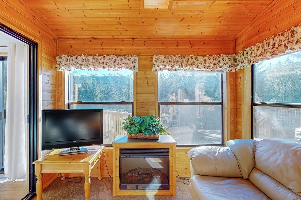 A wooden cabin interior with a television, a fireplace, a sofa, and large windows.