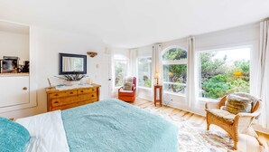 Quarto com cama, cômoda, cadeira e grandes janelas com vista para o verde.