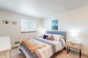 A bedroom with a bed, bedside tables, a window, and a painting on the wall. - Updated condo with river view, convenient location, & private balcony (Seaside)